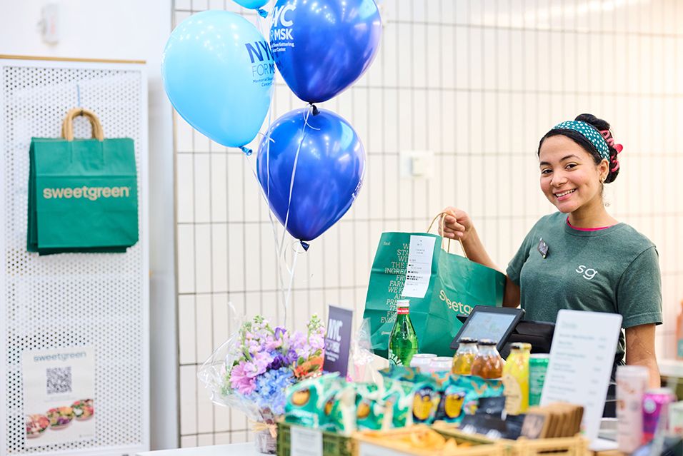 A Sweetgreen employee standing next to blue NYC for MSK balloons holding a green takeout bag.  