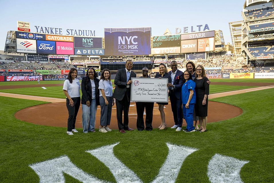 A group of people, including MSK President and CEO Dr. Selwyn M. Vickers, stand behind home plate in Yankee Stadium, holding a large check and smiling. 