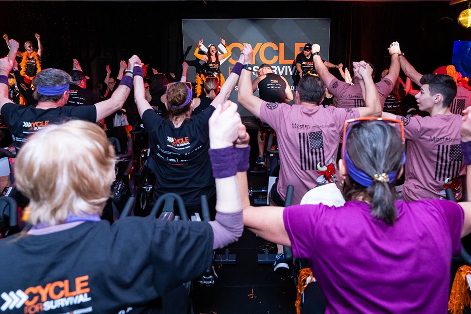 Rows of Cycle for Survival event participants seated on stationary bikes with joined, raised hands, facing a stage.  
