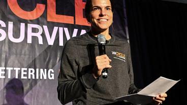 Dr. Diane Reidy-Lagunes, wearing a Cycle for Survival shirt, holds a microphone and smiles. 