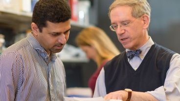 Two men in a discussion, one pointing at papers. Woman working in the background.